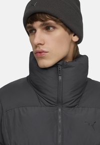 Ung mand iført en mørkegrå beanie og en lynlåset sort pufferjakke, der ser til siden mod en ensfarvet baggrund.