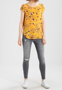Donna che indossa una blusa corta a maniche floreale gialla, jeans skinny grigi strappati e sneakers bianche, in piedi contro uno sfondo semplice.