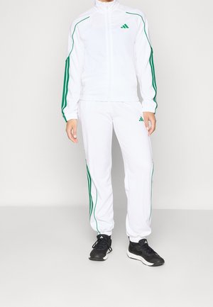 Person iført hvid træningsdragt med grønne striber og logoer, kombineret med sorte og hvide sneakers, stående mod en ensfarvet hvid baggrund.