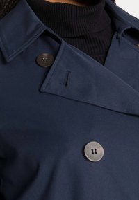 Trench-coat bleu marine avec un col large et des boutons noirs. Texture de tissu lisse avec une coupe sur mesure et un col montant en-dessous.