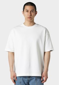 Hvit oversized T-skjorte laget av bomull. Har rund hals, korte ermer, og en liten logo på venstre erme. Glatt tekstur.