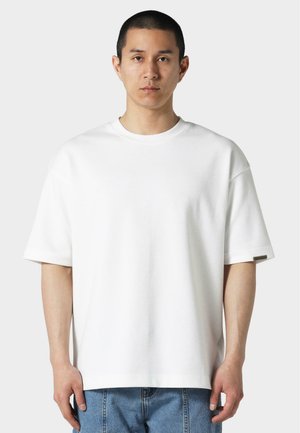 Camiseta blanca de corte oversize hecha de algodón. Presenta un cuello redondo, mangas cortas y un pequeño logo en la manga izquierda. Textura suave.