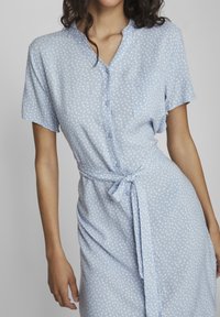 Donna che indossa un vestito corto a maniche corte di colore azzurro chiaro con una fantasia di pois bianchi irregolari, bottoni frontali, tasca sul petto e cintura annodata in vita.