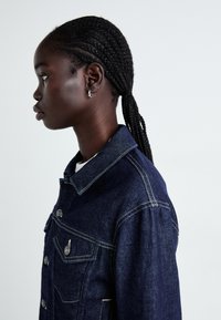 Mörk denimjacka med klassisk krage, silverknappar och framfickor. Slät textur och strukturerad passform. Sidoprofil vy.