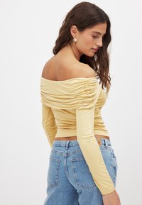 Top z dolgimi rokavi, brez ramenskega dela, nabrana, v svetlo rumeni tkanini, prilegajočega kroja, z gladko teksturo. Uporabljeno z modrimi hlačami visoko pasu.