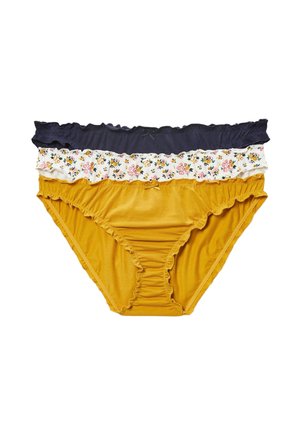 Femme portant un t-shirt blanc uni à manches courtes et un bas de bikini bleu marine, debout devant un fond blanc.