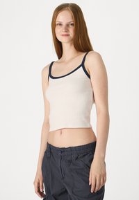 Krēmveida ribota apgrieztā augšdaļa ar tumši zilām maliņām, plānām siksniņām un pieguļošu dizainu, kopā ar tumšiem kravas biksēm.