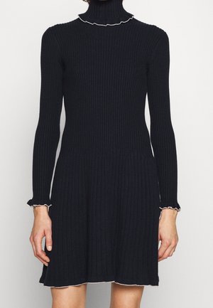 Schwarzes geripptes Strickkleid mit langen Ärmeln, hohem Rüschenkragen und gerüschten Manschetten mit weißem Rand, getragen von einer Person.