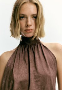 Mlada ženska z kratkimi blond lasmi in pegami, oblečena v brezrokavnik rjave barve z visokim ovratnikom, obrnjena naprej z nevtralnim izrazom na obrazu.