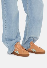 Világoskék díszítéssel ellátott barna nubuk sportcipők, laza szabású világoskék farmernadrággal párosítva. Semleges színű talp és texturált fűzők.