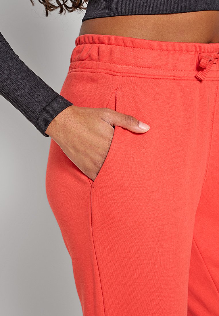 Person, der korallenrote Jogginghose trägt und die Hand in der Seitentasche hat, kombiniert mit einem schwarz langen, cropped Oberteil, das den Bauch zeigt.