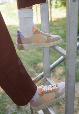 Personne portant des baskets beige, roses et violettes avec des chaussettes blanches, marchant sur une échelle en métal à l'extérieur avec de l'herbe verte en arrière-plan.