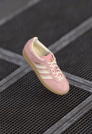 Pink suede sneaker with white three-stripe design, beige accents, and a gum sole. Features laces and branding on the side.