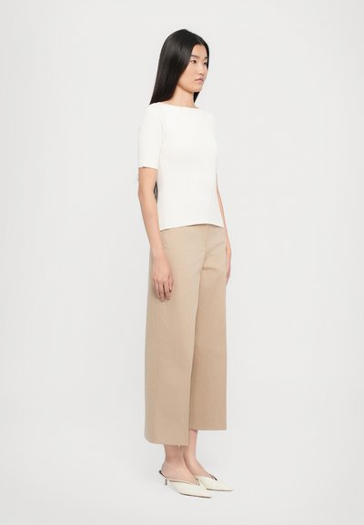 Top bianco a maniche corte a righe abbinato a un pantalone beige a gamba larga. Il modello indossa scarpe slip-on color crema. Design semplice e moderno con colori neutri.