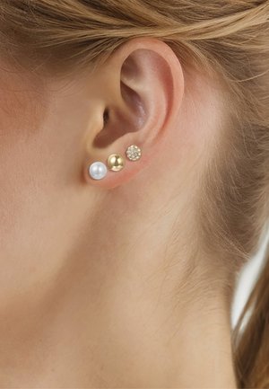 Primo piano di un orecchio sinistro con tre orecchini a perno: perla bianca, sfera d'oro lucida e piccolo cerchio d'oro con cristalli trasparenti.