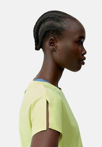 Dzeltena īsām piedurknēm krekls ar sānu šķēlumiem; viegls materiāls. Gluda tekstūra. Slāņots ar zilu apakškrekla, modelis skatīts profilā.