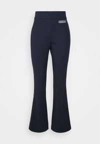 Tmavě modré zvonové kalhoty s žebrovanou texturou, mají vysoký pas a na přední straně nášivku s logem Adidas. Hladký povrch látky.