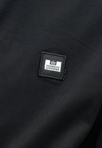 Μαύρο ύφασμα με ένα μικρό τετράγωνο μπαλώμα που απεικονίζει μια κορώνα πάνω από το κείμενο "WEEKEND OFFENDER" με λευκή και μαύρη ραφή.