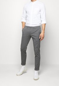 Muž v bílém dlouhém rukávu, šedých slim-fit kalhotách s ohrnutými lemy, bílých ponožkách a bílých teniskách stojící proti bílému pozadí.