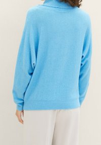 Ljusblå tröja med hög krage, avslappnad passform, ribbade ärmslut och nederkant, samt mjukt, strukturerat tyg. Vågig detalj vid kragkanten.