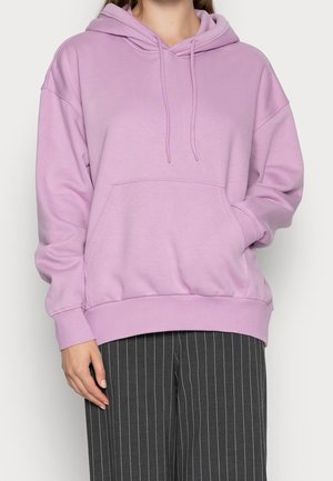 Personne portant un sweat à capuche violet pastel avec poche avant et cordons, associé à un pantalon large gris foncé à fines rayures.