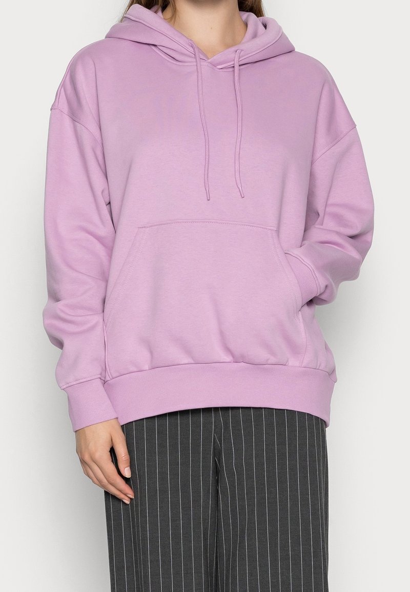 Personne portant un sweat à capuche violet pastel avec poche avant et cordons, associé à un pantalon large gris foncé à fines rayures.