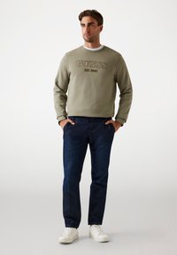 Felpa verde oliva con logo "GUESS EST. 1981", abbinata a pantaloni in denim blu scuro e sneakers bianche. Vestibilità casual e texture morbida.