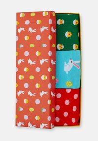 Trois paires de chaussettes colorées dans une boîte orange entrouverte décorée de lapins blancs, d'œufs de Pâques et de pois.