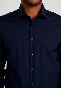 Granatowa koszula z guzikami uszyta z gładkiego materiału, z kołnierzykiem i pięcioma guzikami, z delikatnym wykończeniem na wewnętrznej stronie kołnierza.