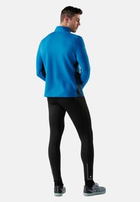 Hombre vestido con una camiseta atlética de manga larga azul, leggings negros con cremalleras y zapatillas grises, de pie de espaldas sobre un fondo blanco.