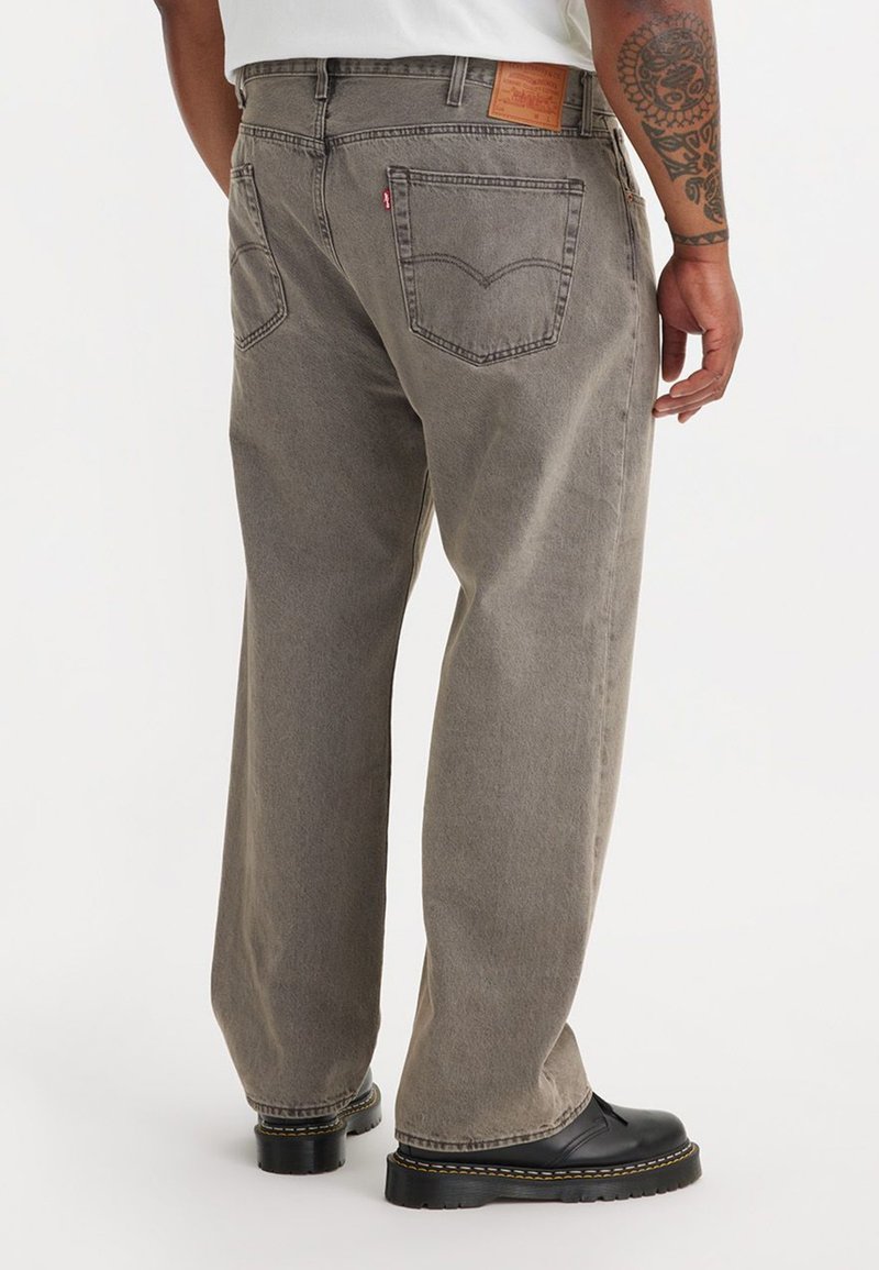 Jean en denim gris avec une coupe décontractée, présentant des poches arrière, une étiquette en cuir marron et des bottes noires avec des coutures jaunes.