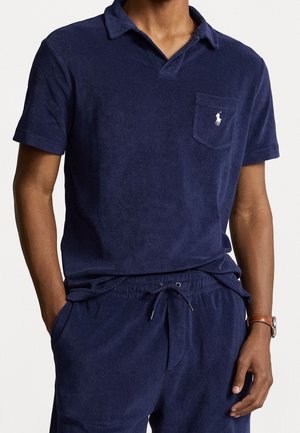Homme portant un polo bleu marine à manches courtes et un short assorti avec cordon, debout les mains dans les poches devant un fond blanc.