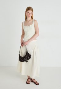 Donna che indossa un top beige senza maniche con bottoni e una gonna lunga, tiene una borsa nera, indossa una collana d'oro, orecchini a cerchio e sandali marroni.
