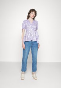 Blusa roxa envolvente com estampa de margaridas brancas, mangas curtas bufantes e um detalhe de laço na cintura, combinada com jeans azul de cintura alta e sapatos claros.
