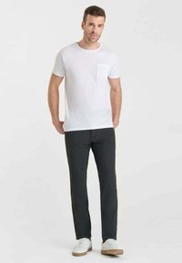 Man die een witte T-shirt met korte mouwen, donkergrijze broek en witte sneakers draagt, staande tegen een eenvoudige witte achtergrond.