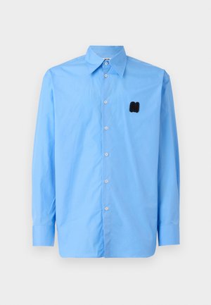 Camisa de manga larga azul claro hecha de tejido suave, con un cuello puntiagudo y un logo bordado en negro en el lado izquierdo del pecho.