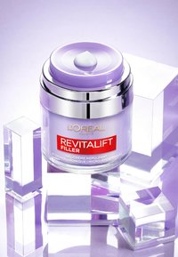 Pot violet avec des accents argentés, contenant un gel crème étiqueté "REVITALIFT FILLER." Présenté sur des blocs acryliques clairs et géométriques.