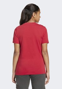 Žena s tmavými vlasmi zviazanými dozadu, oblečená v jednoduchom červenom tričku s krátkym rukávom a šedých šortkách, zobrazená zozadu na bielom pozadí.