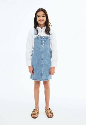 Abito in denim azzurro con bretelle regolabili, bottoni frontali e taglio dritto; abbinato a sandali marroni.