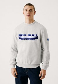 Sweatshirt gris avec des poignets et un ourlet côtelés, arborant un logo "RED BULL" bleu et des rayures horizontales. Porté par un modèle avec les cheveux courts.