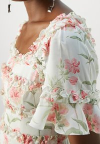 Gros plan d'une robe fleurie avec des fleurs roses et vertes, présentant des bords volantés au niveau de l'encolure, des manches et du corsage.