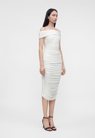 Kvinde iført hvid off-shoulder ruched midi-kjole og sorte ankelstroppet høje hæle, stående mod en ensartet lys baggrund.