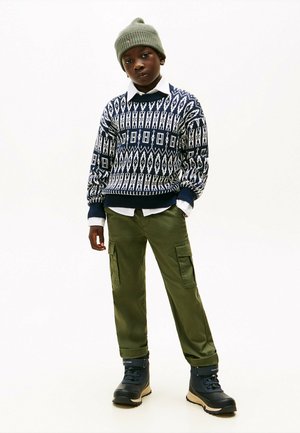 Pull en tricot avec des motifs géométriques bleu marine et blancs, associé à un pantalon cargo olive et des bottes noires. Un bonnet vert complète la tenue.