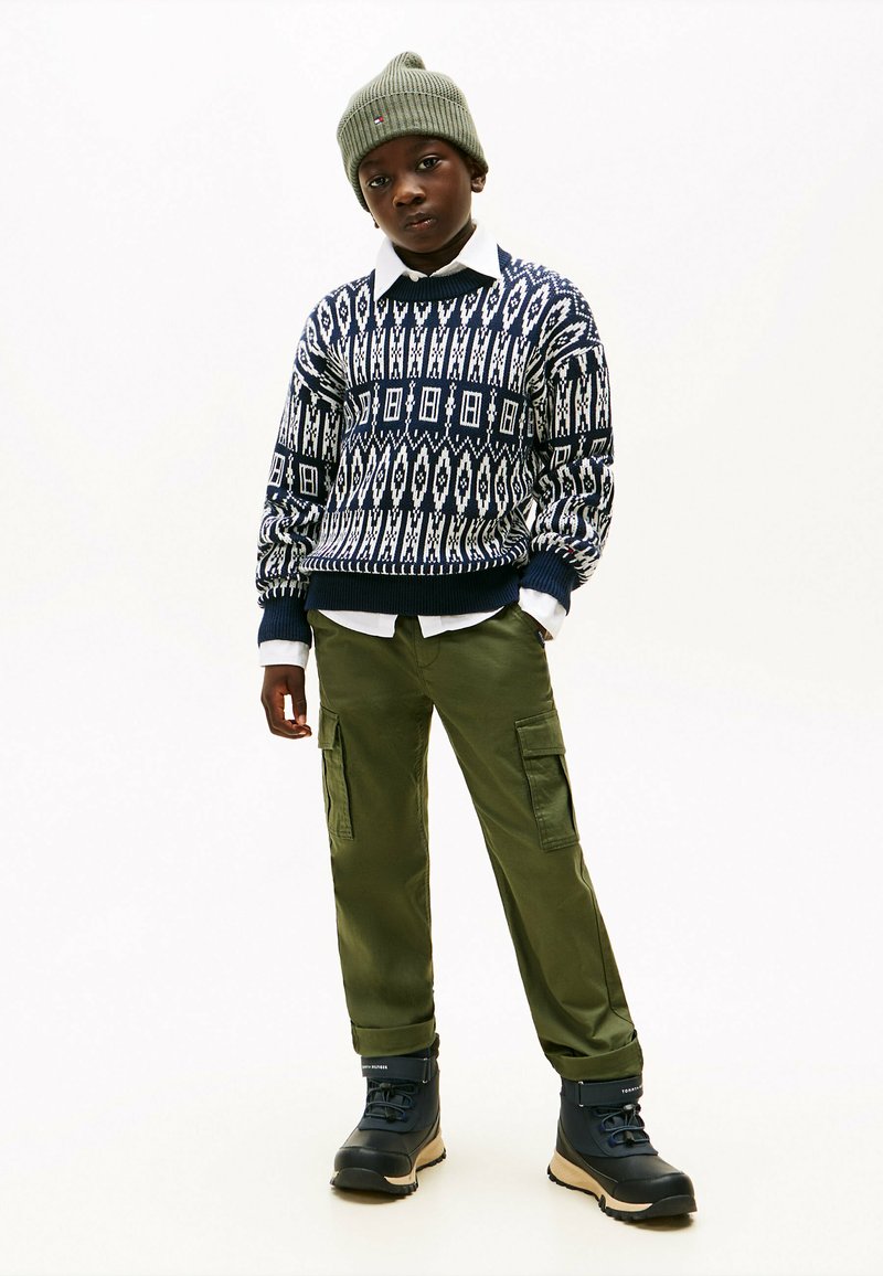 Strickpullover mit navyblauen und weißen geometrischen Mustern, kombiniert mit olivfarbenen Cargo-Hosen und schwarzen Stiefeln. Eine grüne Mütze rundet das Outfit ab.