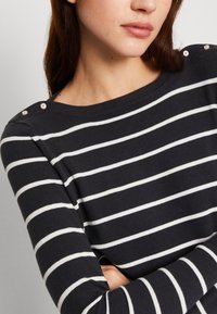 Marinblå randig tröja med vita horisontella linjer, tillverkad av mjukt material, med dekorativa knappar på axeln och en bred halsringning.