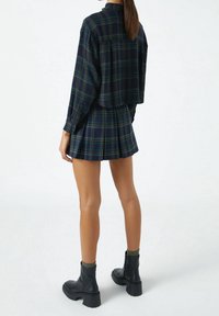 Camicia a quadri navy tagliata, con linee verde e giallo, abbinata a una gonna plissè coordinata. Stivali neri alla caviglia con suole spesse.