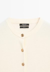 Gilet en laine de couleur blanc cassé avec un col côtelé. Il dispose de deux boutons dorés sur le devant pour la fermeture. Étiquette de marque à l'intérieur du col.