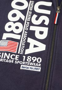 Marinblå zip-up sweatshirt med stor vit text, en amerikansk flagga-patch och broderade detaljer. Inkluderar en dragkedja med en färgad accent.