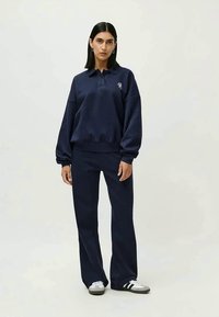 Conjunto de sweatshirt e calças azul marinho, com colarinho, punhos canelados e detalhe de logotipo. O tecido parece suave e texturado. Ténis complementam o visual.