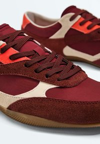 Pair of maroon and beige suede sneakers with brown laces and orange accents, on a white background.